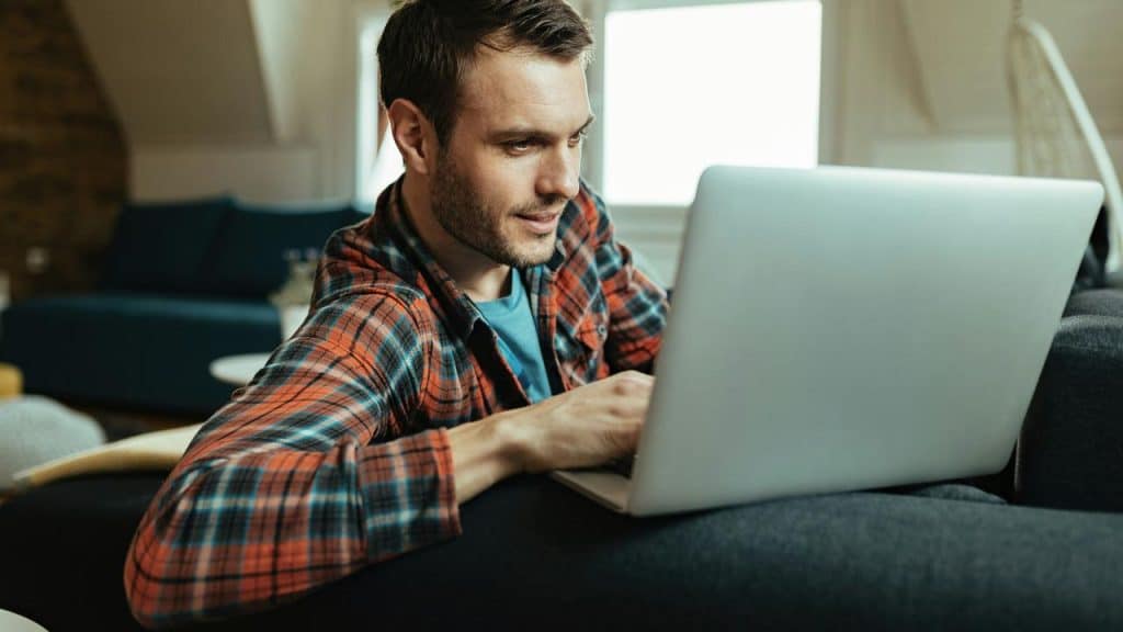 A man in a plaid shirt uses a laptop on a couch at home.