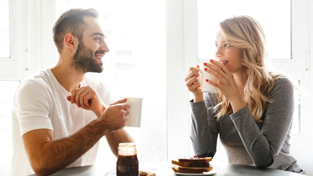A man and woman sit at a table drinking from mugs and looking at each other.
