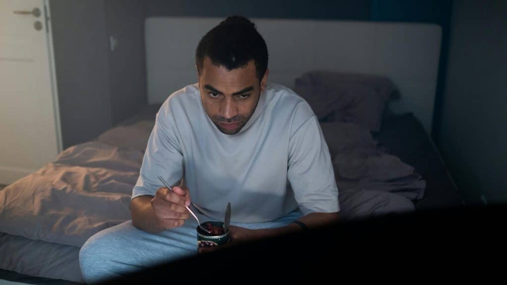A man sits on a bed in a dark room, eating from a small tin.