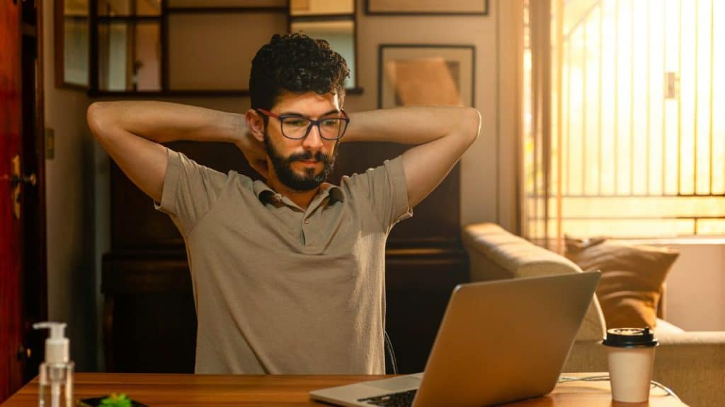A man with glasses and a beard sits at a laptop with hands behind head.