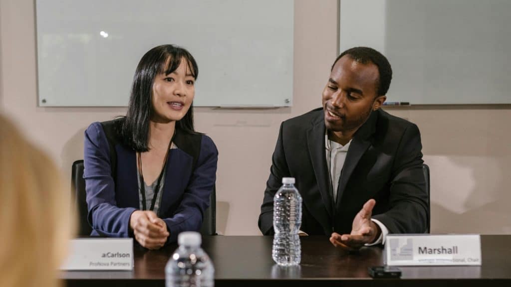 A woman and a man sit at a table with nameplates and talk during a meeting.