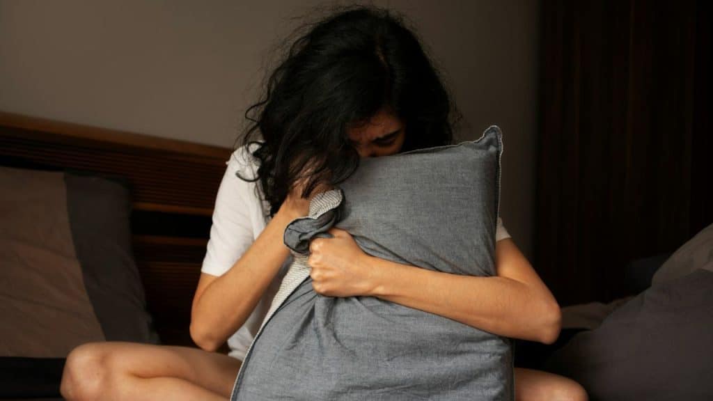 A person with dark curly hair sits on a bed and tightly hugs a gray pillow.