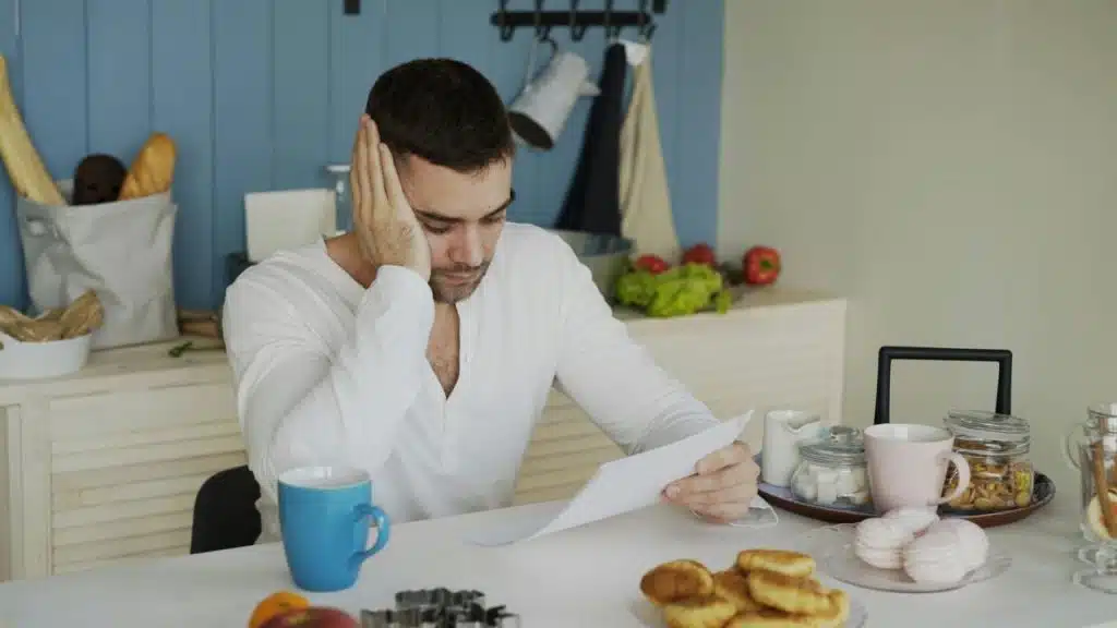 A man in a white shirt sits at a table reading a paper with his head down.