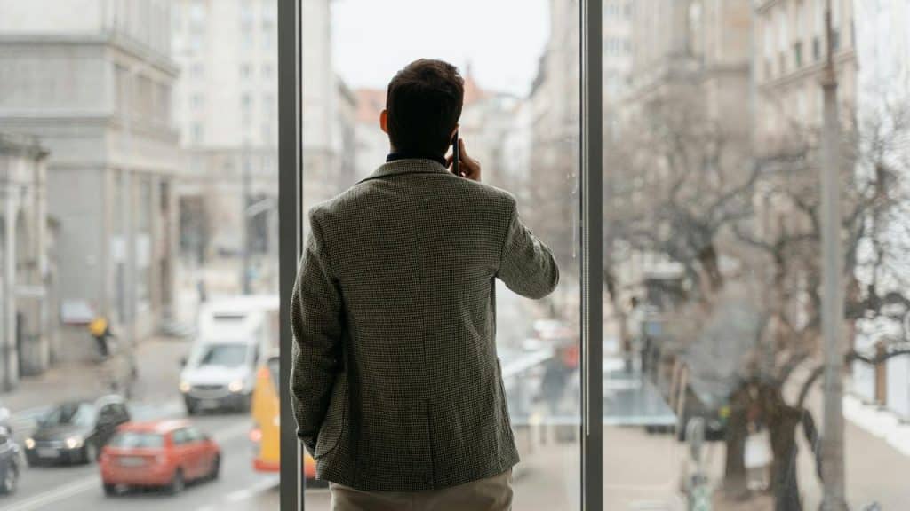 A man standing by a large window talking on his phone while looking at the city.