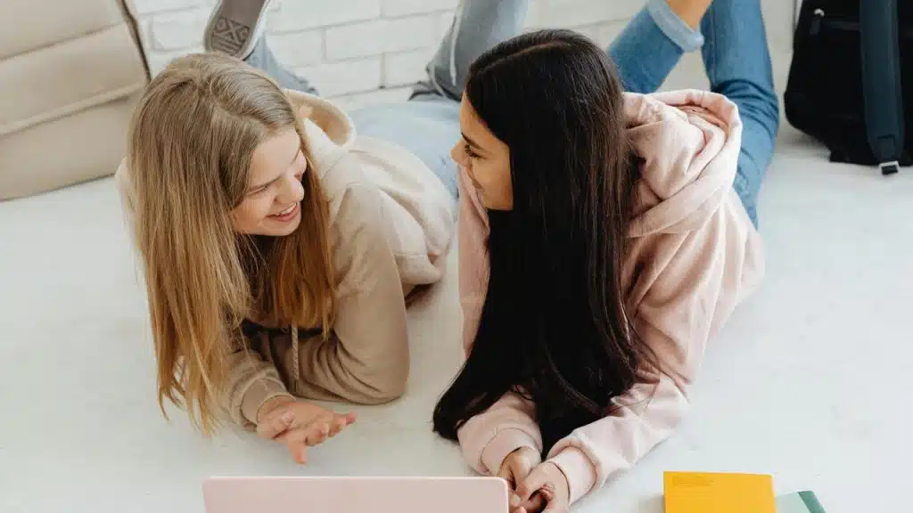 A pair of young women lying on the floor and talking while using a laptop.