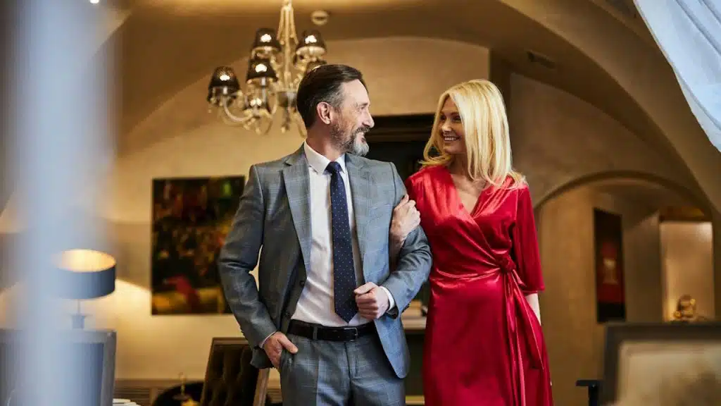 A well-dressed couple walking arm-in-arm indoors, smiling at each other in a lit room with a chandelier and decorative interior in the background.