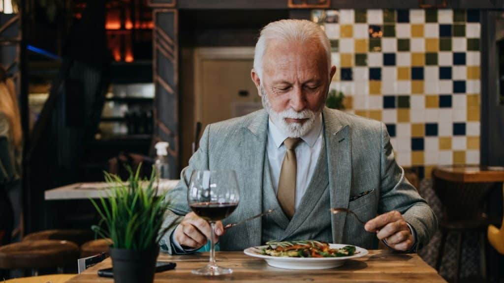 An older man in a grey suit eats a meal alone at a restaurant table.