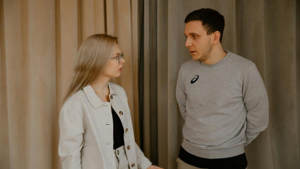 A woman with glasses and a man in a gray sweatshirt stand talking near curtains.