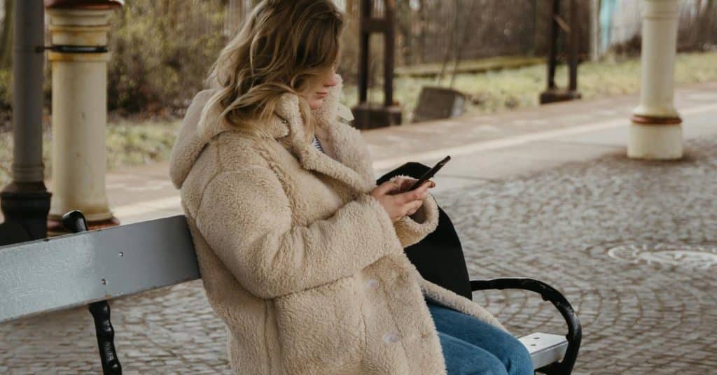 A person in a warm coat sits on a bench and looks at a phone.