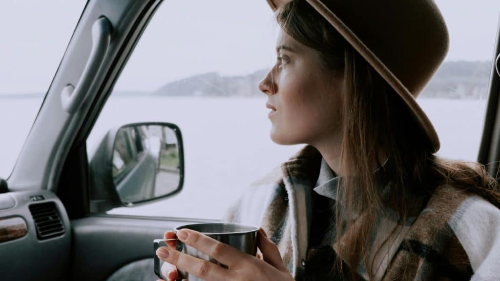 A woman sitting in a car, holding a cup and looking out the window thoughtfully.