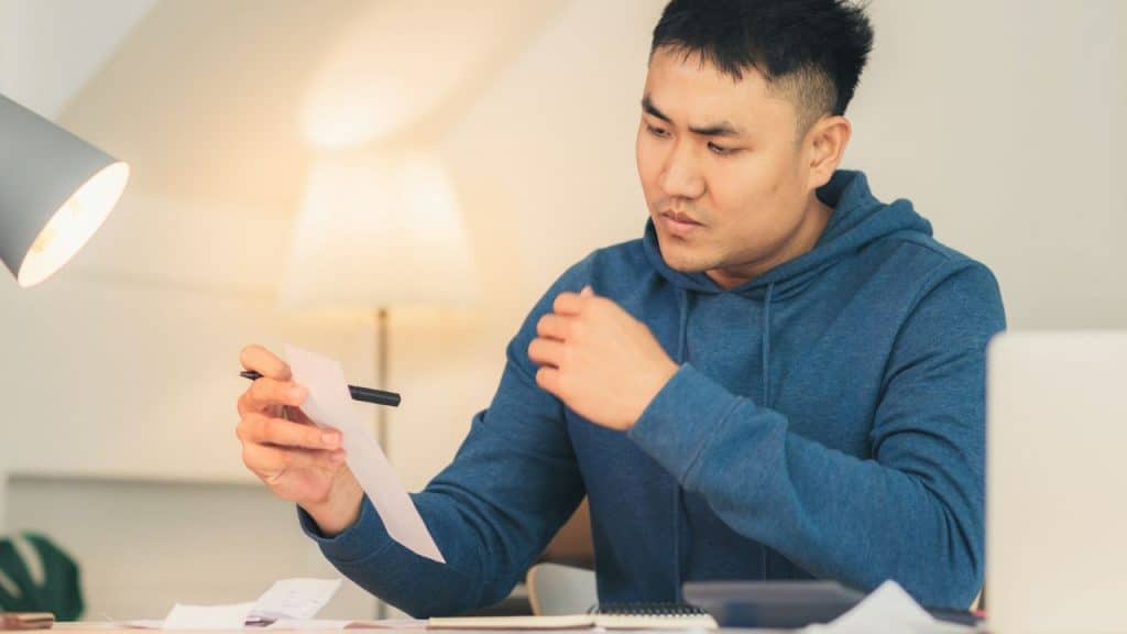 A man in a blue hoodie sits at a desk looking at a white piece of paper.