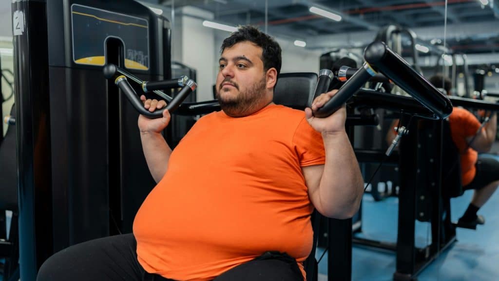 A man in an orange shirt uses a shoulder press machine in a brightly lit gym.