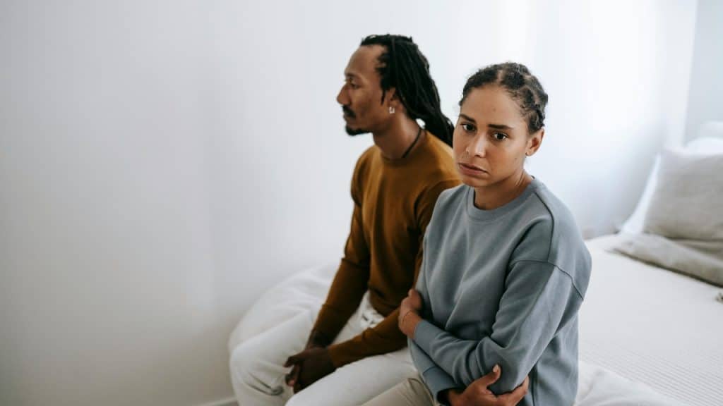 A woman with crossed arms sits on a bed while a man looks away from her.