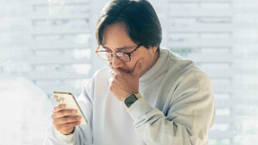 A man with glasses and a light sweater holds a smartphone while touching his chin outdoors.