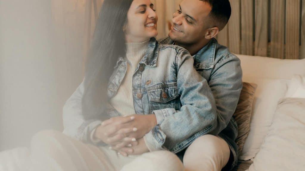 A couple cuddling on a bed and smiling at each other.