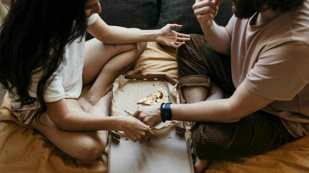A couple sitting on a bed sharing a pizza from an open box.