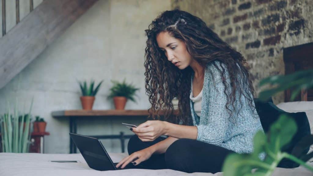 A woman with long curly hair holds a credit card while looking at a laptop screen.