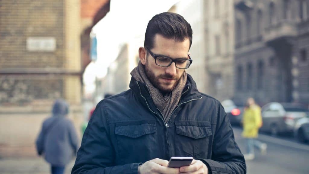A man with glasses and a scarf looks down at his phone on a street.