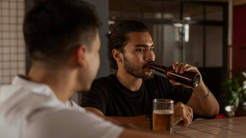A man drinking beer while sitting at a bar with another person nearby.