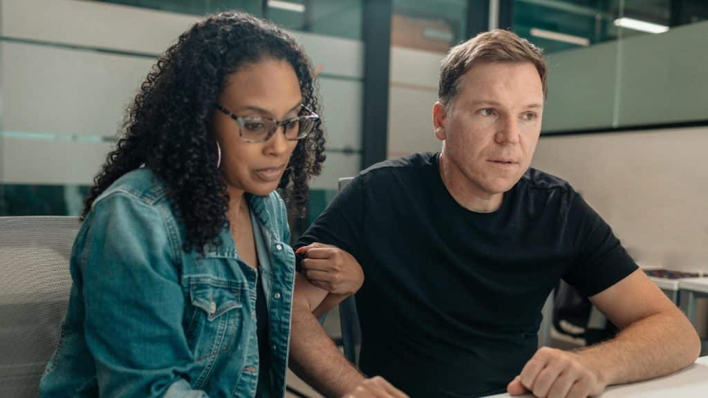 A man and woman sit side-by-side in an office setting, looking at a screen.