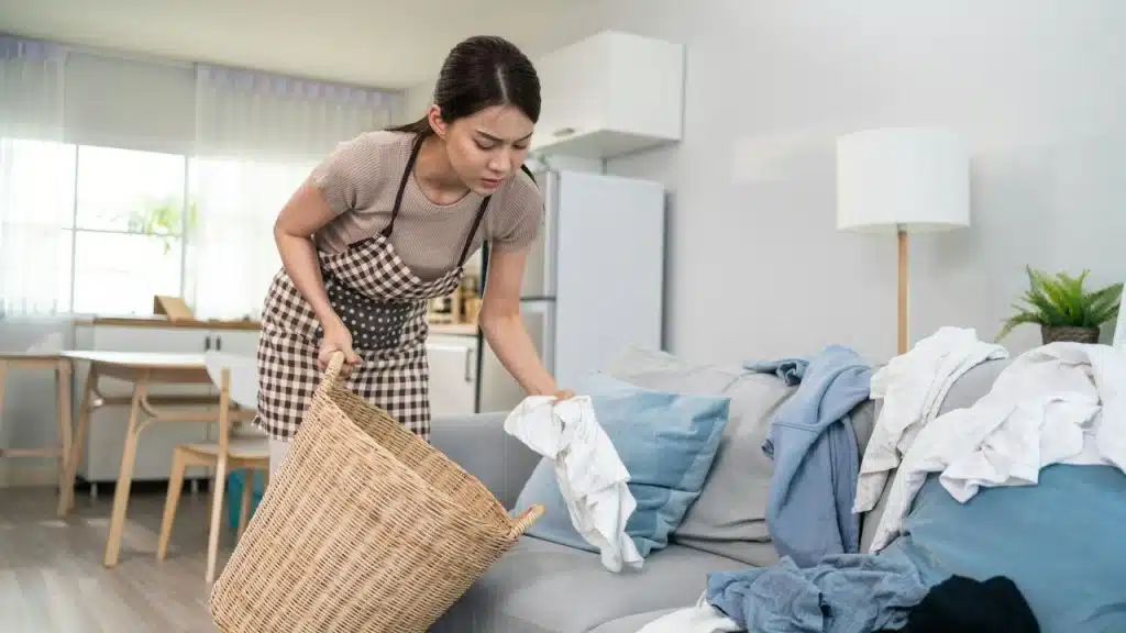 A woman in a checkered apron places white clothing into a large woven laundry basket.