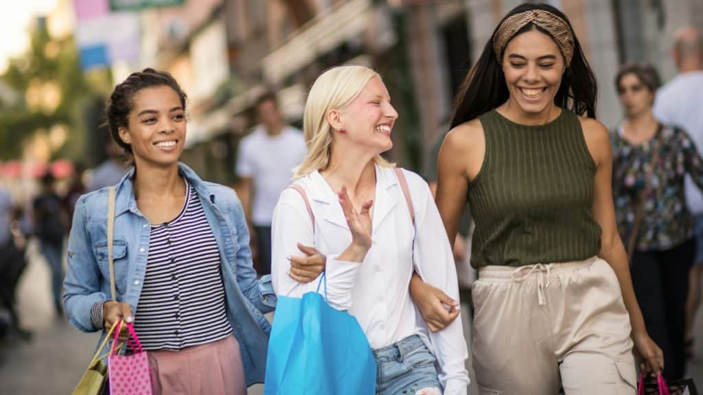 Three women walk down a city street together while smiling and holding colorful shopping bags.