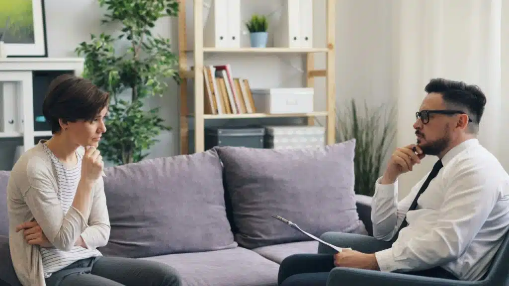A woman sits on a couch across from a man holding a clipboard in an office.