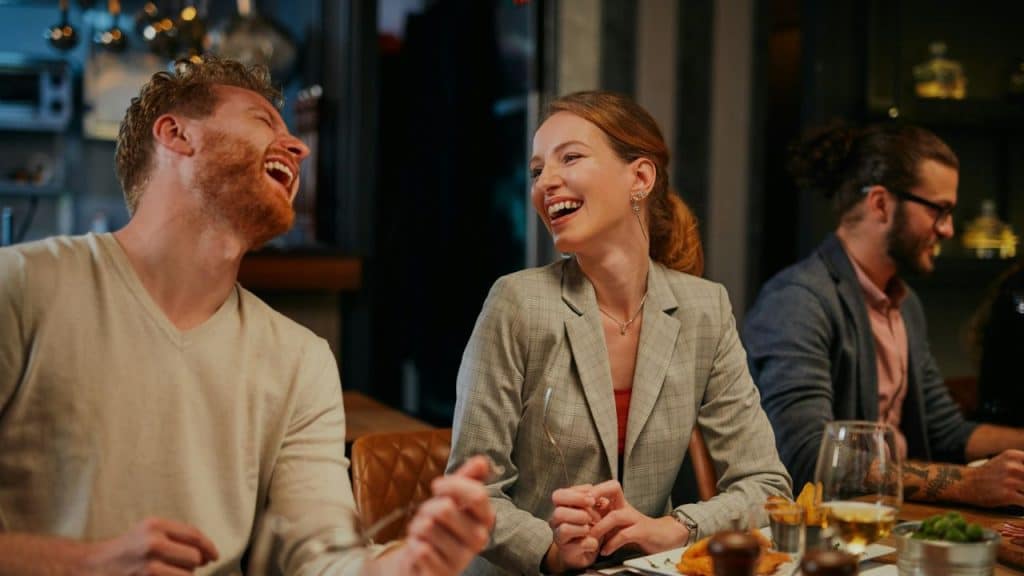 A man and woman laugh together while sitting at a table in a crowded restaurant.