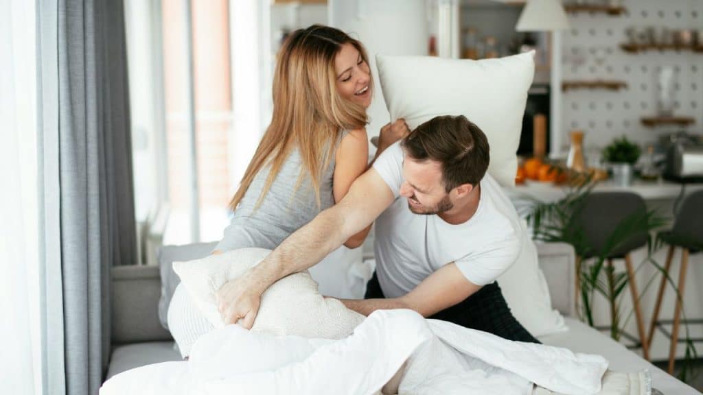 A laughing man and woman engage in a playful pillow fight on a bed together.