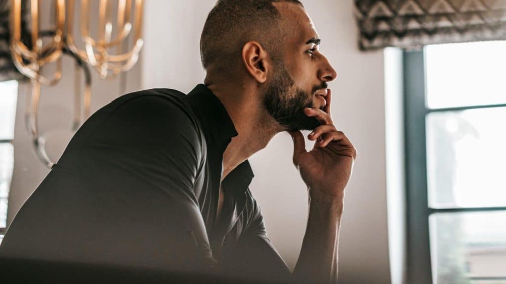 A thoughtful man in profile resting his chin on his hand by a window.
