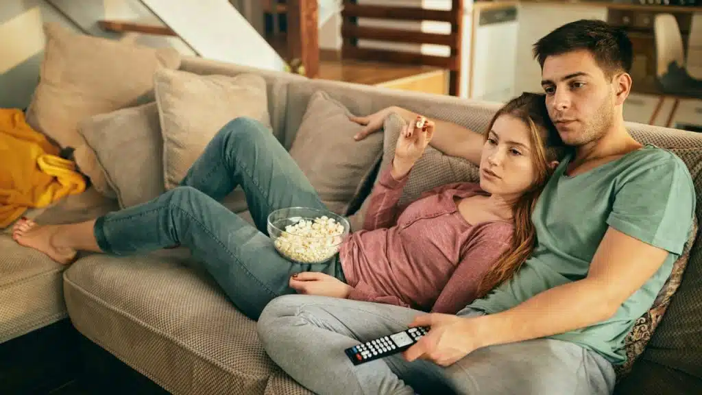A man and woman lean against each other on a couch while holding a remote.