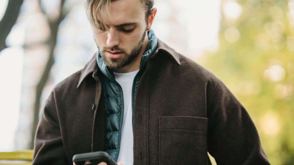 A man with a beard and brown jacket looks down at a smartphone while outdoors.