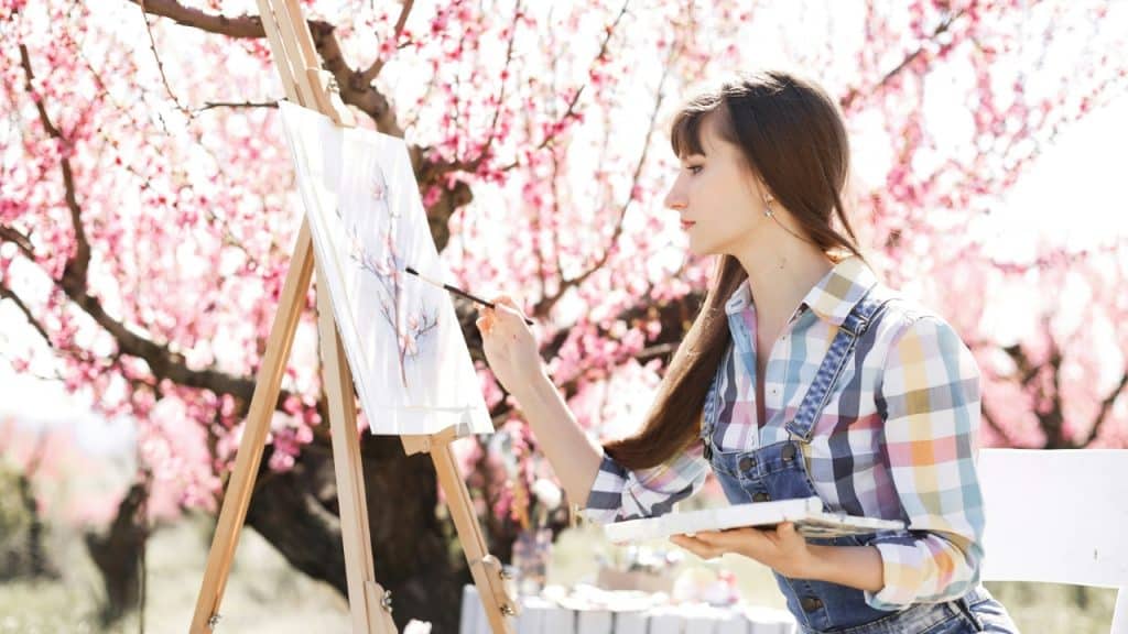 A woman in a plaid shirt and overalls paints a canvas under blossoming trees.