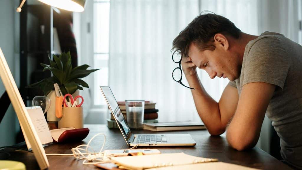 A man leaning over a desk with a laptop, holding his glasses and appearing tired while working.