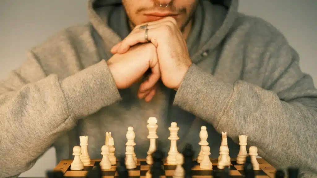 A person in a grey hoodie sits behind a chessboard with white and black pieces.