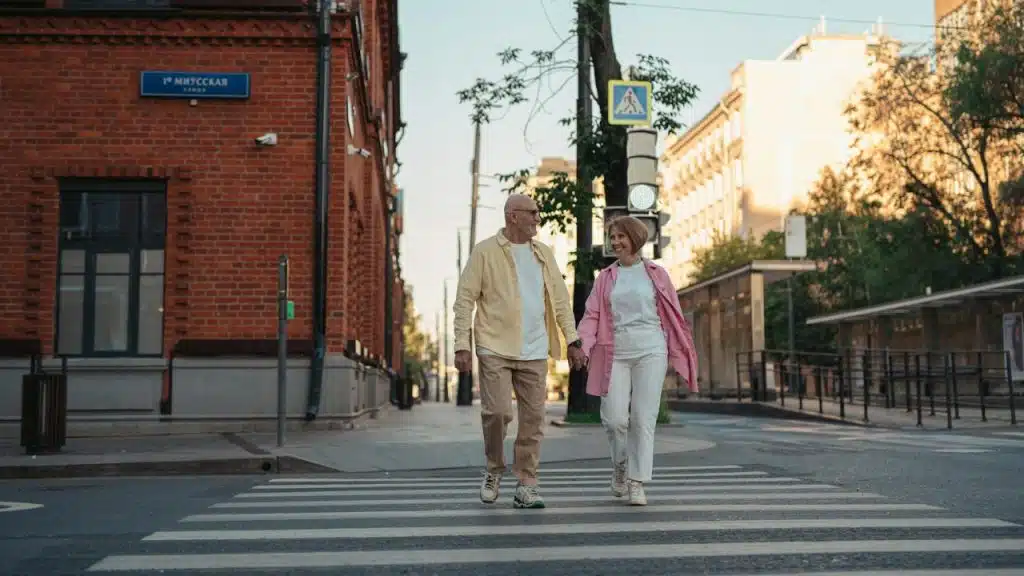 An older man and woman hold hands while walking across a city street crosswalk.
