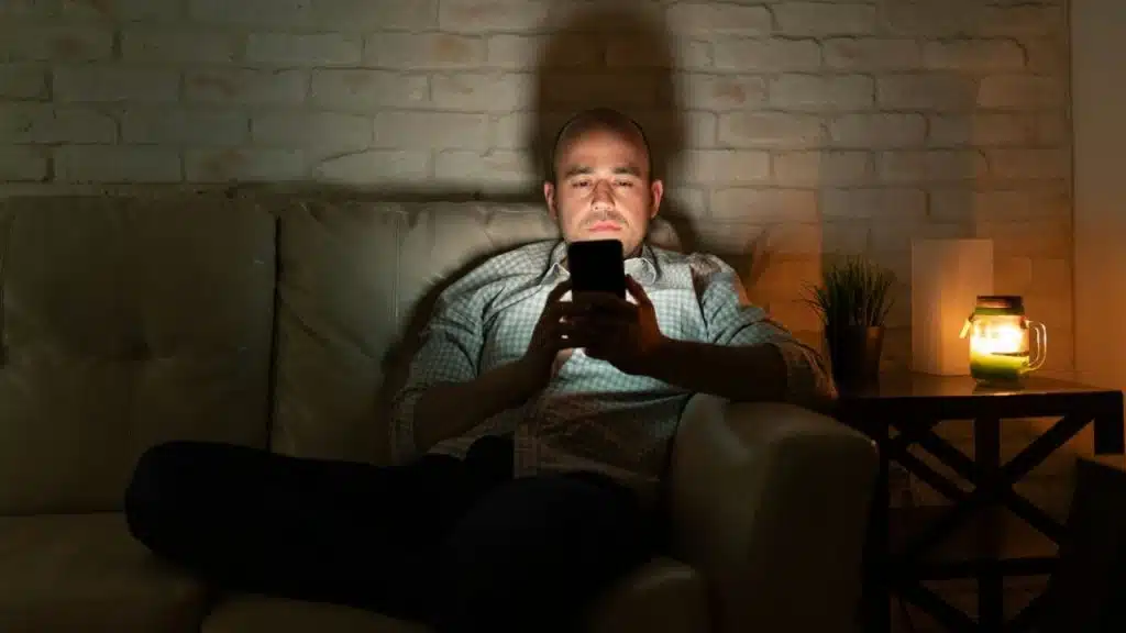 A man sits on a couch in a dark room looking at his glowing phone screen.