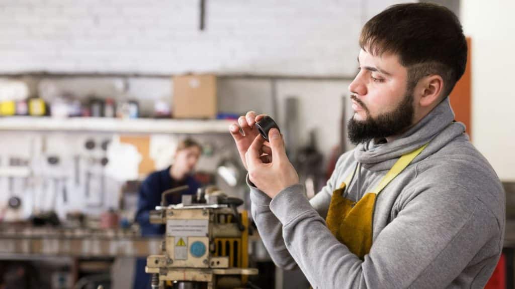 A bearded man in an apron examines a small circular object in a workshop setting.
