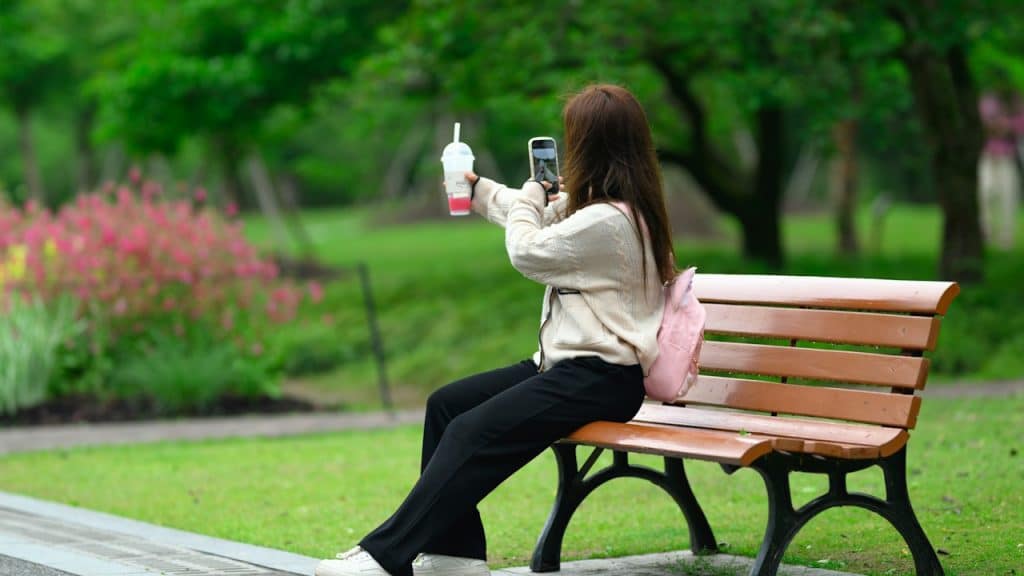 A woman taking a picture of her drink while sitting on a bench in the park.