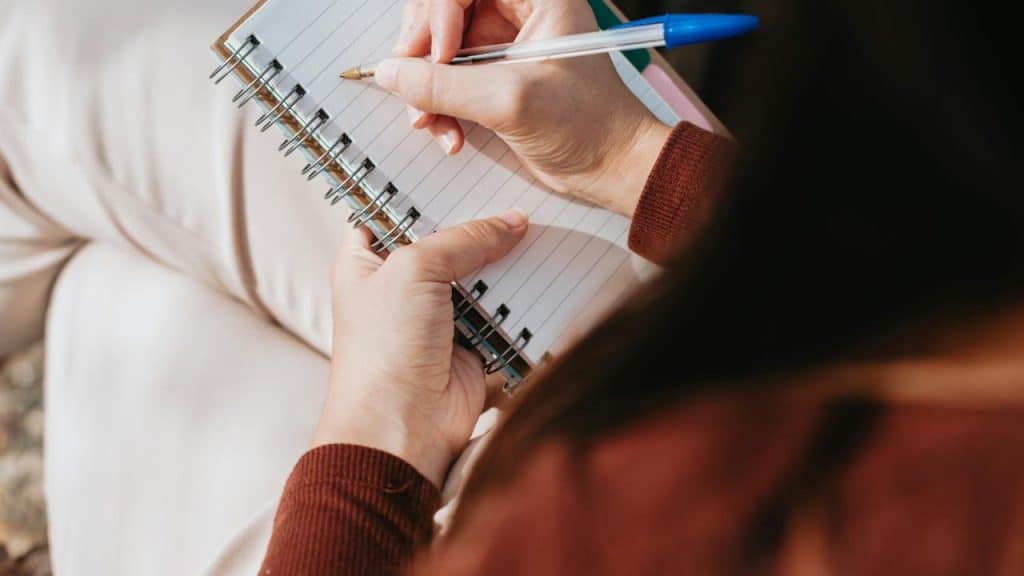A person writing in a spiral notebook with a pen.