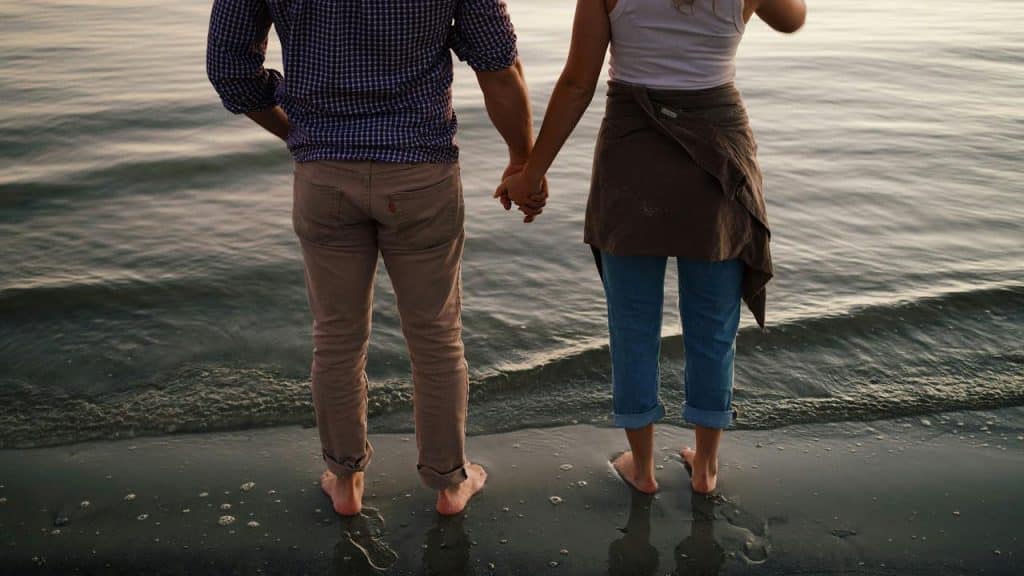 A couple holding hands while standing barefoot at the water’s edge.