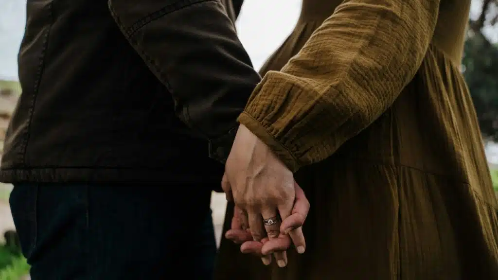 A close-up shot shows two people standing side-by-side outdoors, holding hands firmly.