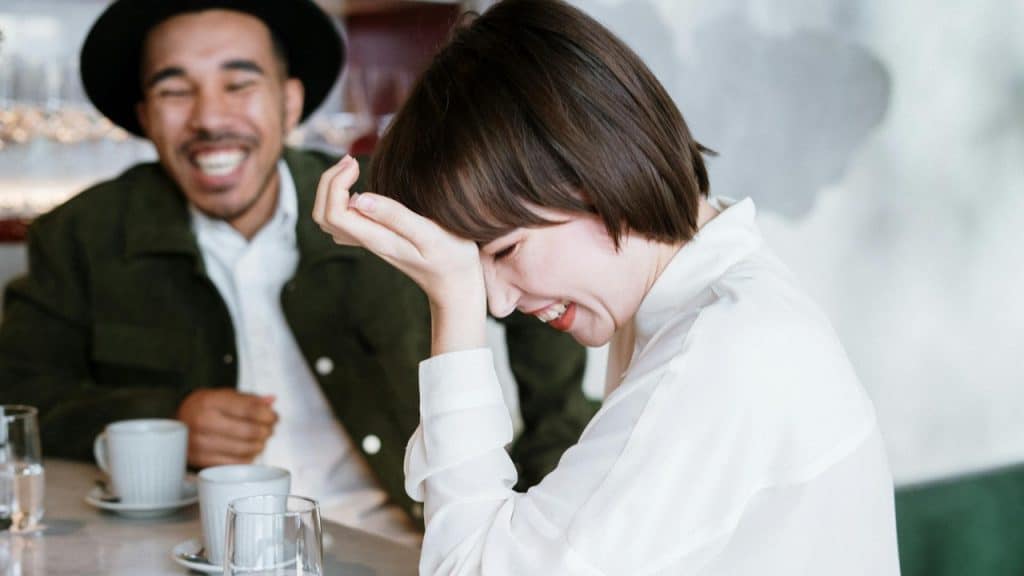 A woman with short hair laughs with her hand on her forehead near a man.