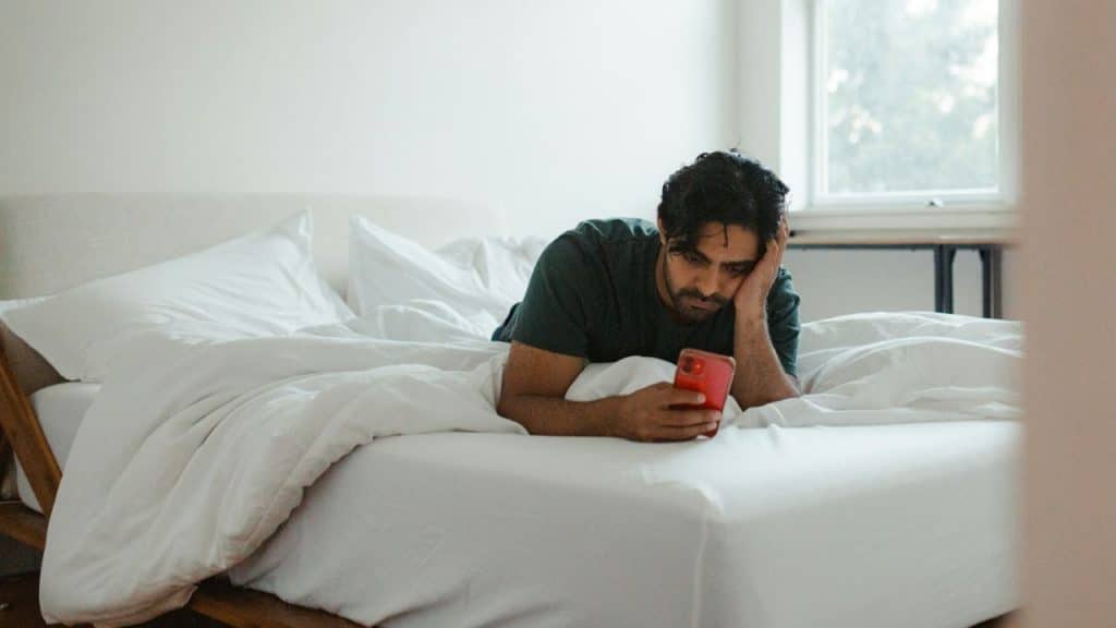 A man lies on a bed with white linens while looking at a red smartphone.