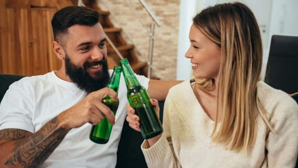 A bearded man and a woman toast with green glass bottles while sitting on a sofa.