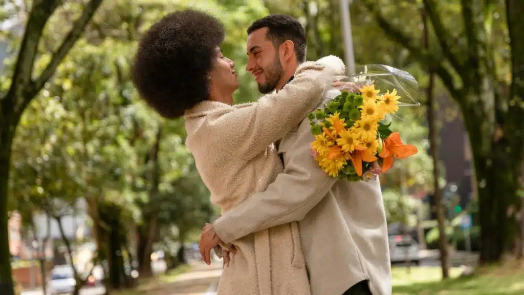 A man and woman embrace outdoors while the man holds a bouquet of yellow flowers.