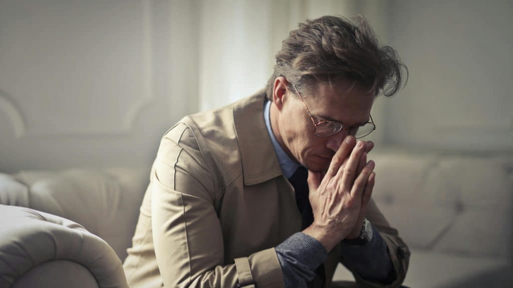 A man in a trench coat sits with his head down and hands pressed together.