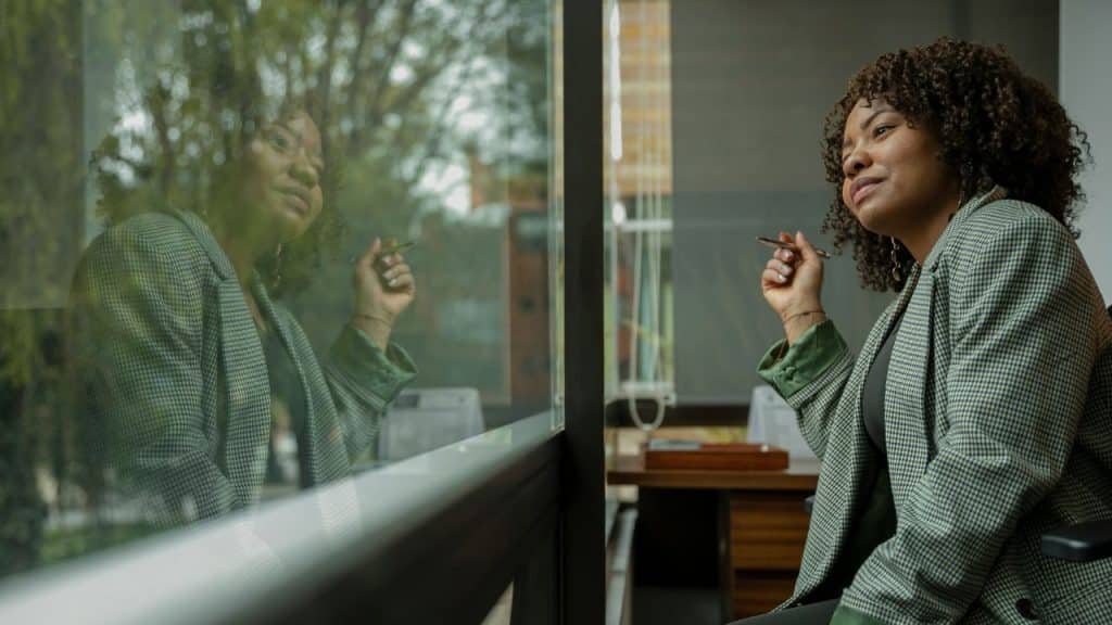 A woman in a blazer holds a pen and looks out of a large glass window.
