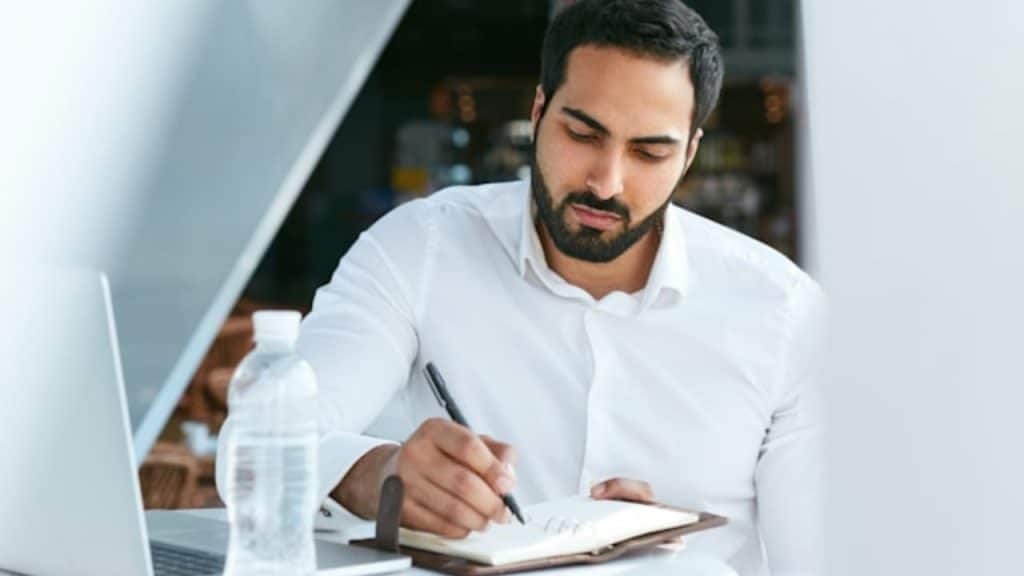 A man with a beard wearing a white shirt writing in a notebook at a desk.