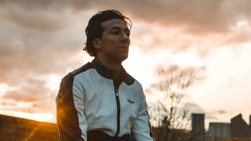 A man in a white and black jacket looks forward against a bright sunset background.